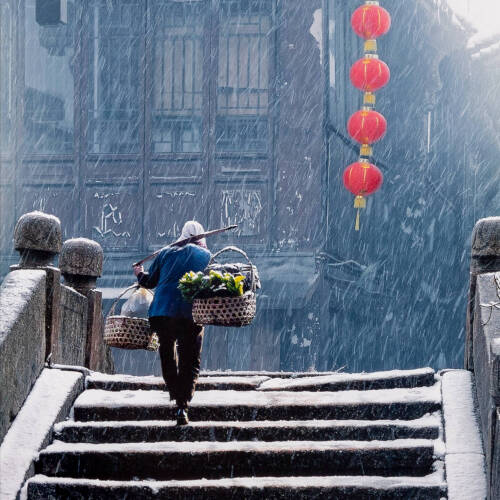 Color photograph of a man carrying to baskets on his shoulders walking up a set of stairs in the snow.